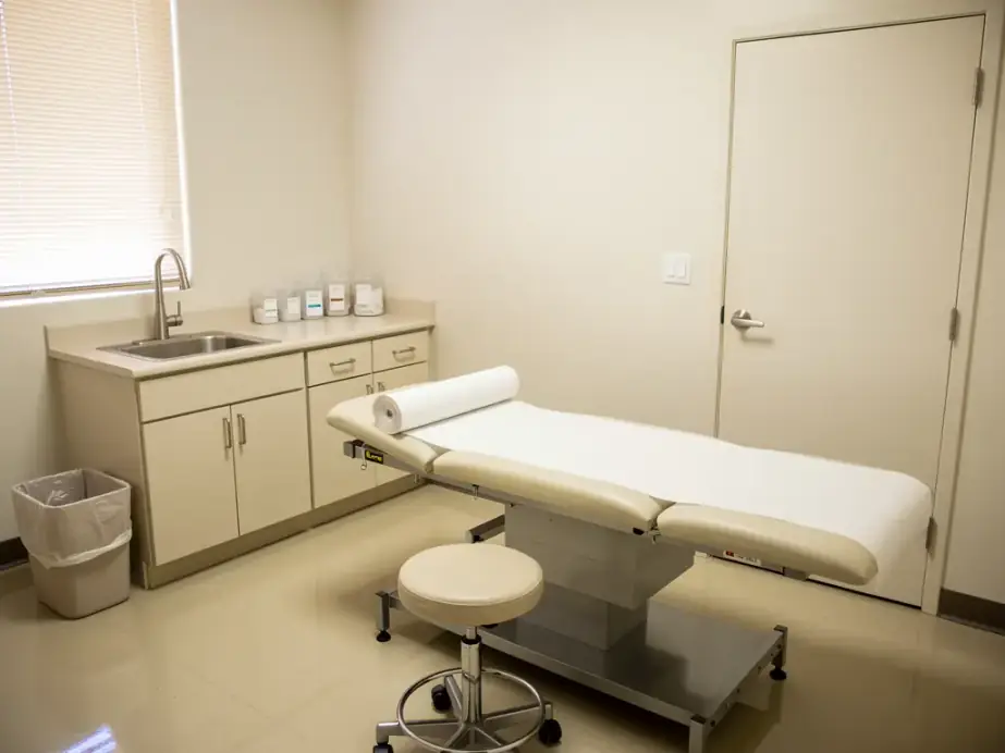 A clean medical exam room: exam table with a paper roll, a stainless sink, a lever-handle door and a lined waste bin